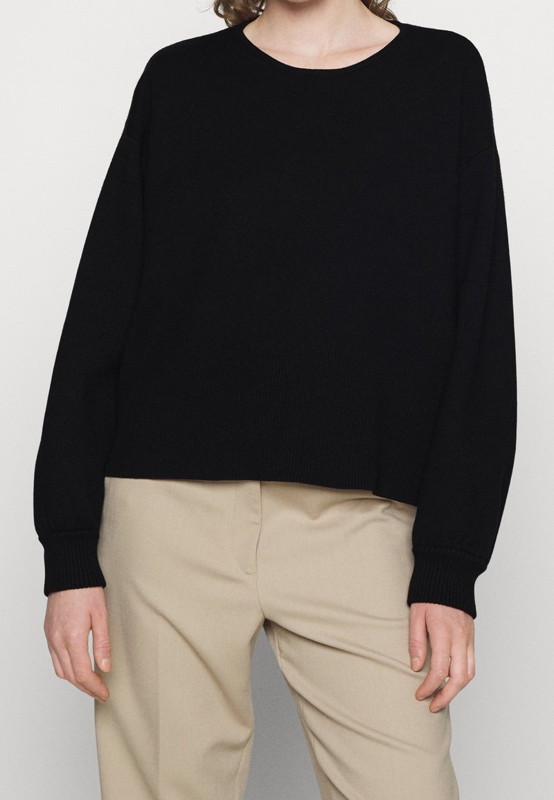 Pull en tricot noir avec un col rond et des coutures d'épaules tombantes. Texture côtelée aux poignets et à l'ourlet, associé à un pantalon beige.