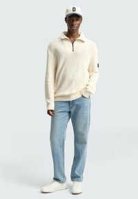Cremfarbener Strickpullover mit einem halben Reißverschluss, kombiniert mit hellblauen Jeans und weißen Sneakers. Die passende Mütze hat einen dunklen Schirm und ein Logo.