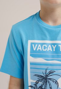 Ljusblå bomullströja med tryck som har texten "VACAY TIME", horisontella ränder och palmer i svart. Rund halsringning.