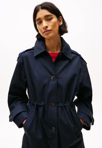 Cappotto trench blu navy con vita cintrata, grandi bottoni e colletto classico. Dotato di tasche laterali e maniche arrotolate.