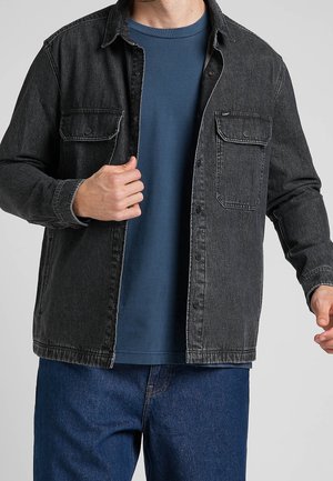 Man wearing unbuttoned black denim jacket over blue shirt and blue jeans, adjusting the jacket with left hand.