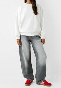 Persona che indossa una felpa bianca, jeans grigi larghi e sbiaditi, e sneakers rosse con suole in gomma, che si trova in piedi contro uno sfondo bianco semplice.