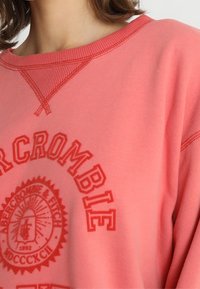 Koralfarvet sweatshirt med ribbet krave og manchetter. Har præget logo og tekstdesign i en matchende farve. Blødt, struktureret materiale.