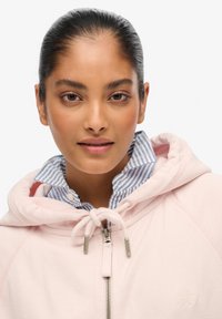 Roze zip-up hoodie met een trekkoord, zachte textuur en logo. Gelaagd over een blauw-wit gestreept overhemd met kraag.