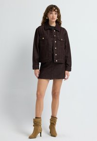 Giacca di jeans marrone con tasche frontali, bottoni argentati e una gonna in jeans marrone abbinata con orlo sfilacciato. Abbinata a stivaletti alla caviglia color sabbia.