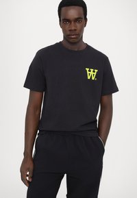 Zwarte katoenen t-shirt met een klein felgeel logo op de borst. Het model draagt bijpassende zwarte broek met een relaxte fit.