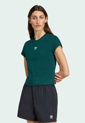 Camiseta de algodón verde con mangas cortas y cuello redondo, con un logo blanco de Adidas centrado en el pecho. Combinada con pantalones cortos negros.
