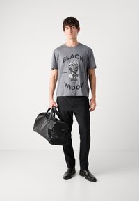 Grå grafisk t-shirt med texten "BLACK WIDOW", svarta byxor och en stor svart läder gympapåse, bärs av en manlig modell som står mot en enkel bakgrund.