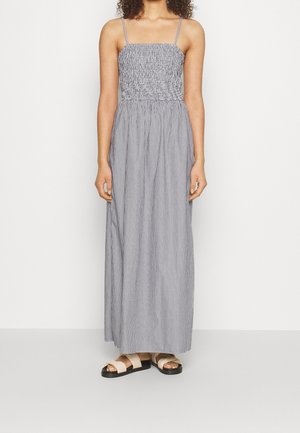 Kobieta ubrana w długą, bezrękawnikową, szarą sukienkę w paski na cienkich ramiączkach, stojąca na tle jednolitego białego tła, ubrana w beżowe sandały.