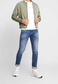 Kurtka bomberka w oliwkowej zieleni z prążkowanymi mankietami, biała koszulka z grafiką, niebieskie dżinsy i białe trampki z czarnymi akcentami.