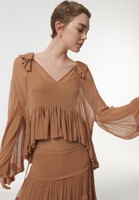 Haut transparent marron avec des détails volants, décolleté en V et accents en nœud sur les manches. Tissu léger qui s'écoule librement, superposé à une jupe assortie.