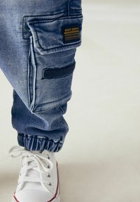 Denim joggerji svetlo modre barve z elastičnimi manšetami, stranskimi žepi in črnim tekstilnim žepkom; v kombinaciji z belimi supergami z rdečimi dodatki.