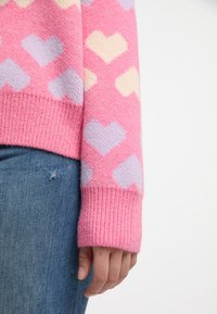 Maglione lavorato a maglia con motivi a cuore rosa, lilla e crema. Il maglione presenta un polsino a coste e una finitura morbida e testurizzata. Indossato con jeans blu.