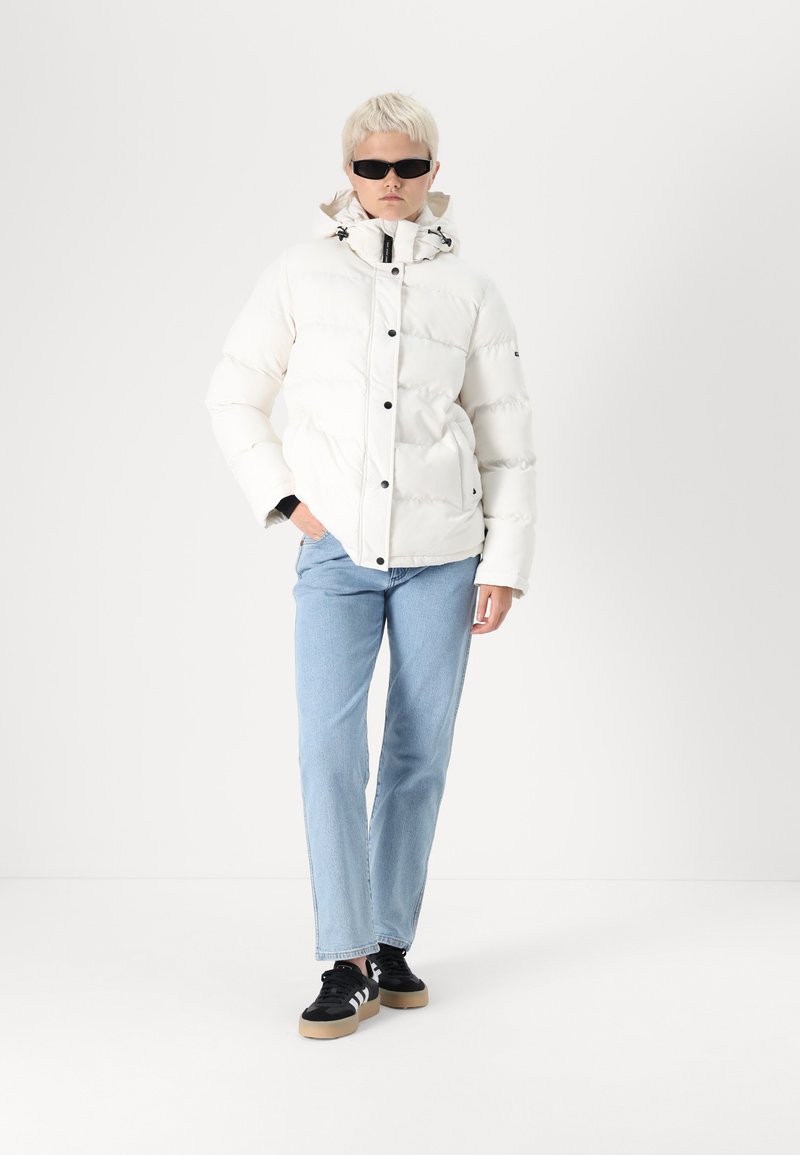 Weißer Puffer-Jacke mit hohem Kragen, schwarzen Knöpfen und Seitentaschen, getragen über blauem Jeans und schwarzen Sneakern. Stilisiert mit Sonnenbrille.