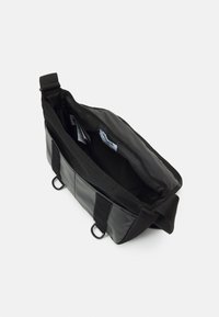 Svart axelremsväska med texturerad yta i nylonmaterial, justerbar rem och kardborrebandstängning; öppen vy visar innerficka och varumärkesetikett.