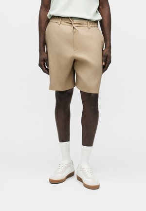 Mann trägt beige Kordelzug-Shorts, weiße Knöchelsocken und weiße Turnschuhe mit braunen Sohlen, steht vor einem einfarbigen Hintergrund.