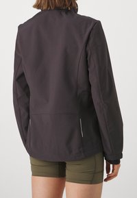 Veste technique noire avec un col montant, une coupe ajustée et des accents réfléchissants ; associée à un short vert olive. Texture mate.