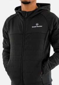 Giacca nera con zip, caratterizzata da una texture imbottita, vestibilità slim e logo Sergio Tacchini sul lato sinistro del petto. Design con cappuccio e tasche laterali.