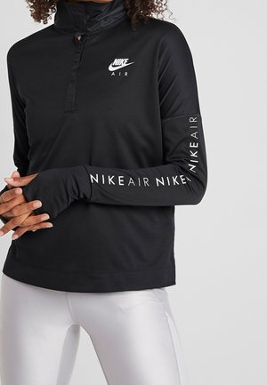 Personne portant un haut à manches longues noir Nike Air avec des trous pour les pouces et des leggings blancs sur un fond clair uni.