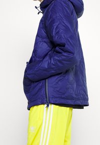 Mörkblå dunjacka med huva, lättviktig material, dragkedjefickor och elastiska ärmmuddar, parat med ljusgula träningsoverallsbyxor.