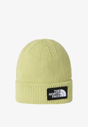 Gorro de punto en amarillo claro con textura acanalada y puño doblado. Presenta un parche de logo negro con texto blanco y detalles de diseño.