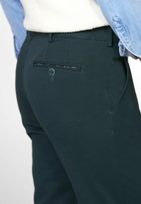 Donkergroene op maat gemaakte broek van een gestructureerde stof, met een achterzak voorzien van een patroon voering en een blauwe knoopaccent.