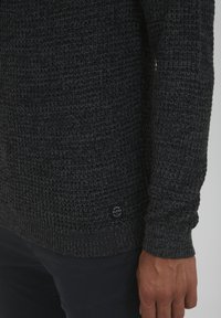 Dunkelgrauer Strickpullover mit einem strukturierten Stoff, taillierten Ärmeln und einem kleinen runden Logo-Patch near dem Saum.