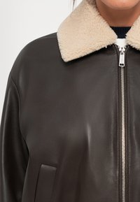 Dark brown leather jacket with cream sherpa collar and silver front zipper worn over a black and white top.