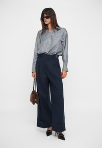 Chemise rayée bleu et blanc à boutons, coupe ample, portée avec un pantalon large à taille haute bleu marine et des escarpins noirs à bout pointu. Sac à main marron.