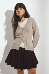 Bežový pletený cardigan s výstrihom do V, piatimi gombíkmi a dvoma prednými vreckami. Nosí sa cez bielu košeľu a v páre s čiernou skladanou sukňou.