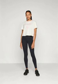 Zkrácené krémové tričko s růžovým logem "GUESS", spárované s černými legínami a černými teniskami. Modelka stojí s rukama v bocích.