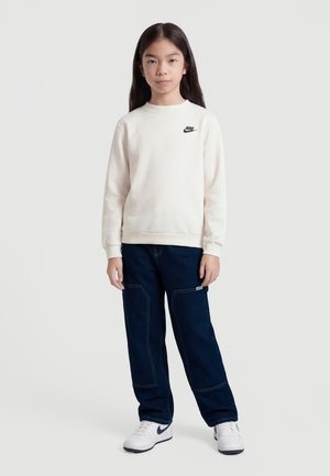 Meisje staand, gekleed in een witte Nike-trui, donkerblauwe broek en witte sneakers met zwarte Nike-logo's, tegen een effen lichte achtergrond.