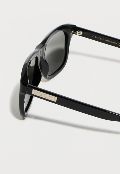 Μαύρα ορθογώνια γυαλιά ηλίου Gucci με σκούρους φακούς, με πλακέτα λογότυπου Gucci σε χρυσό στον βραχίονα και κείμενο "Κατασκευασμένο στην Ιταλία".