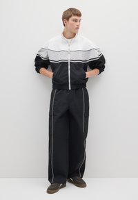 Zwart-witte windjack met ritssluiting, horizontale strepen en elastische manchetten, gecombineerd met zwarte cargobroek met witte biesdetails.