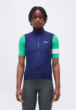 Persona que lleva un chaleco de ciclismo sin mangas azul marino sobre una camiseta de manga corta verde con un brazalete blanco, y pantalones cortos de ciclismo oscuros.