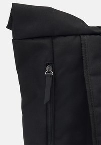 Sac à dos en tissu noir avec une poche zippée verticale et une languette en cuir sur le devant, montrant le bord supérieur roulé et la boucle de poignée.