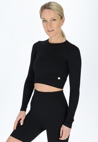 Sort ribbet crop top med lange ærmer, der har en rund halsudskæring og guld logoaccent. Sat sammen med højhuskede sorte shorts.