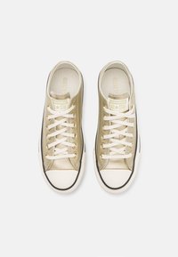 Zlaté plátěné tenisky s bílými tkaničkami, gumovou špičkou a černým lemováním. Obsahují štítek s logem na jazyku a mají hladký povrch.