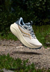 Kevyt beige juoksukenkä, jossa on teksturoitu verkkopäällinen, harmaat yksityiskohdat, oranssit tehosteet ja pehmustettu pohja, leijumassa soratien yllä.