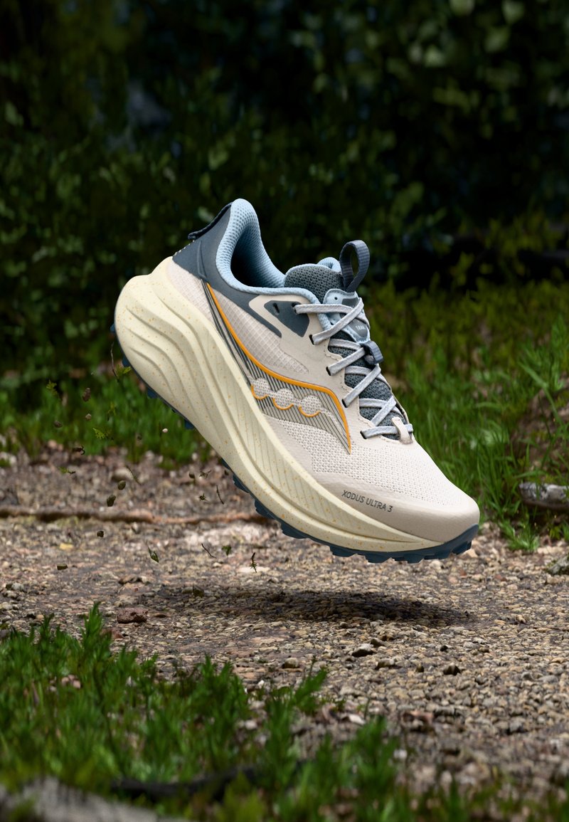 Kevyt beige juoksukenkä, jossa on teksturoitu verkkopäällinen, harmaat yksityiskohdat, oranssit tehosteet ja pehmustettu pohja, leijumassa soratien yllä.