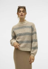 Beige gebreide trui met gestreepte patronen in donkerblauw. Hoge hals, ruime pasvorm en ribbelmansjetten. Gedragen met een bijpassende beige rok.