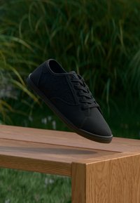 Schwarze Schnürsneaker mit einer strukturierten Oberseite, runder Spitze und Gummisohle, platziert auf einem Holztisch vor unscharfem grünem Gras.