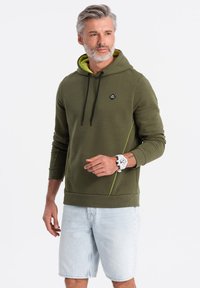 Sudadera con capucha de color verde oliva con cordones negros, forro verde lima y detalles con cremallera, combinada con pantalones cortos de mezclilla claros.