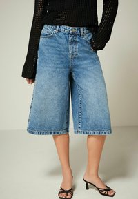 Culottes en denim bleu clair à taille haute, avec un ourlet effiloché et des poches avant. Associées à des sandales à talons noires à lanières.