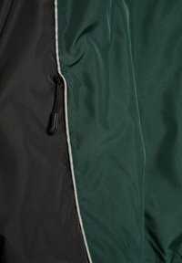 Nahaufnahme einer grün-schwarzen Jacke mit einem vertikalen Reißverschluss und weißer Einfassung entlang der Naht zwischen den beiden Farben.