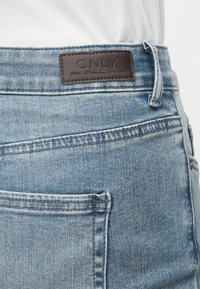 Jean en denim bleu clair avec une étiquette en cuir marron portant la mention « ONLY » à la taille, porté avec une chemise blanche rentrée.
