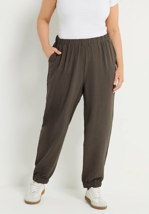 Osoba ubrana w ciemnobrązowe spodnie dresowe z elastycznym pasem i bocznymi kieszeniami, białe trampki oraz włożoną do środka białą koszulę z krótkim rękawem.