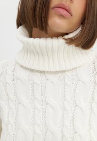 Pull en tricot blanc avec un col côtelé haut, motif en maille torsadée, texture douce et coupe ajustée.