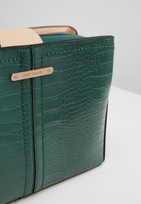 Borsa in finta pelle verde scuro con texture a coccodrillo, dotata di rifiniture beige e di una placca con logo "RIVER ISLAND" in tono oro.