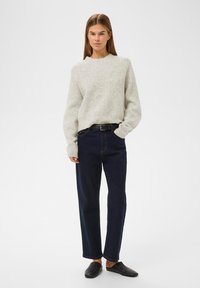 Žena stojící s rukou v kapse, oblečená v světle šedém pleteném svetru, tmavě modrých džínách s černým páskem a černých nazouvacích botách na bílém pozadí.